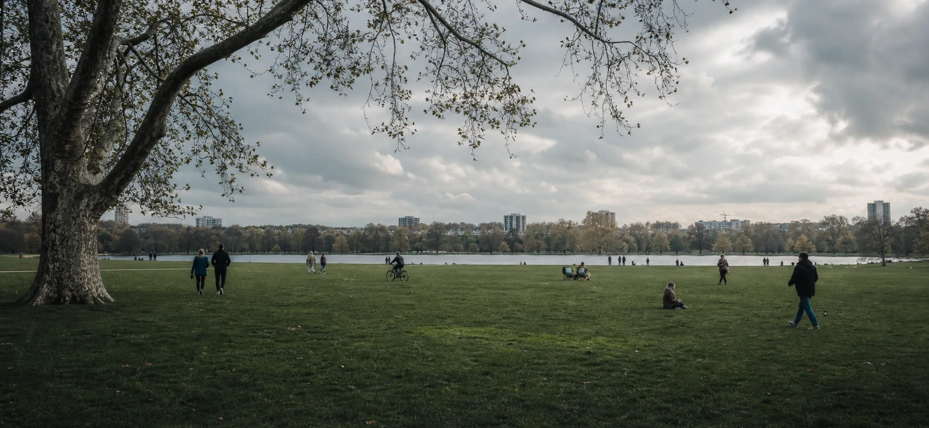 Weite Rasenfläche im Hyde Park mit Wasser, altem Baumbestand, vereinzelten Menschen und ferner Stadt